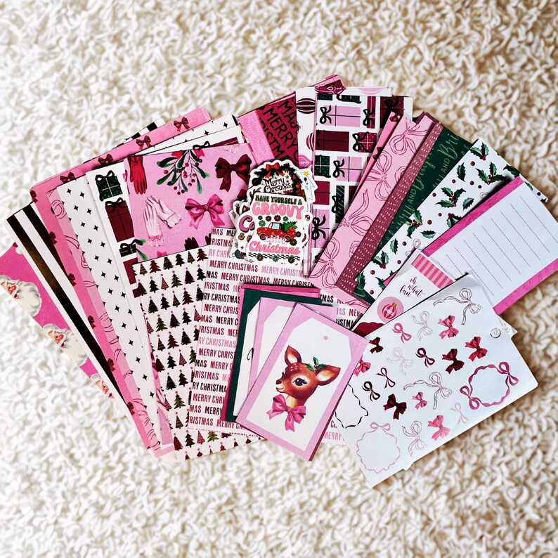 May include: A selection of pink and white Christmas paper ephemera. The collection includes patterned papers, gift tags with a deer illustration, stickers, and notepads. Some items feature bows, Christmas trees, and the text "Merry Christmas."