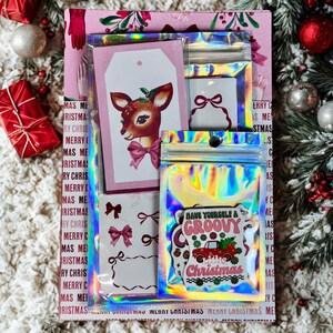May include: A festive Christmas gift set with a pink and white patterned box. It contains gift tags with a deer illustration, stickers, and holographic pouches. One pouch reads "Have Yourself a Groovy Little Christmas."