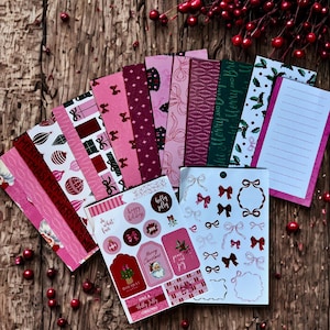 May include: A selection of pink and red Christmas stationery and stickers. The stationery includes patterned paper, lined notepaper, and stickers with bows, festive greetings, and Santa Claus illustrations. The items are arranged on a wooden surface with red berries.