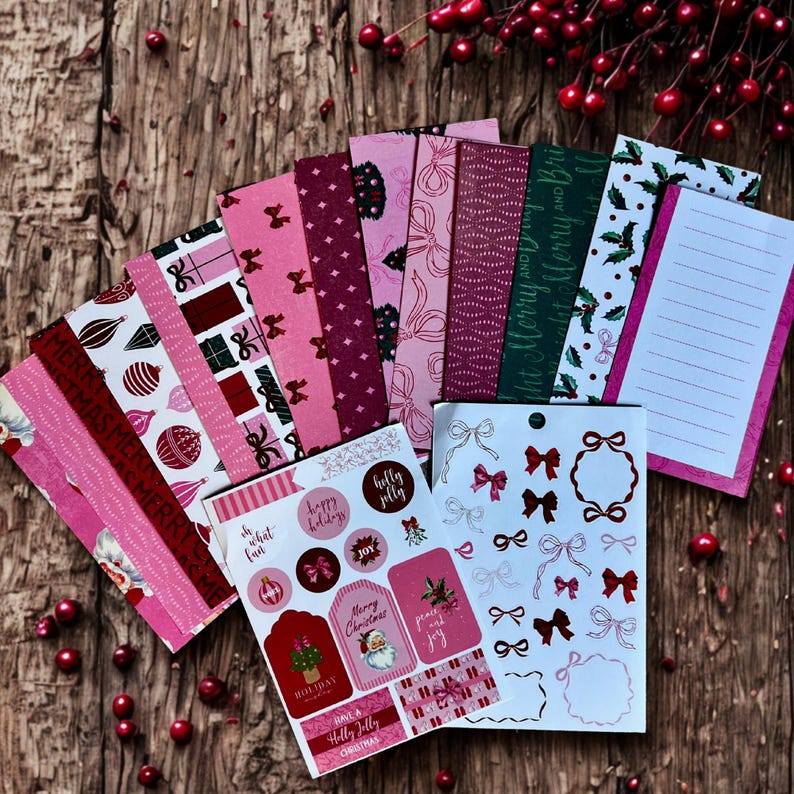 May include: A selection of pink and red Christmas stationery and stickers. The stationery includes patterned paper, lined notepaper, and stickers with bows, festive greetings, and Santa Claus illustrations. The items are arranged on a wooden surface with red berries.