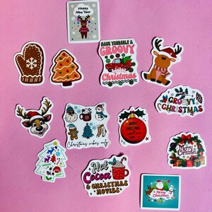 May include: A selection of Christmas stickers featuring festive designs. Includes reindeer, Christmas trees, and gingerbread men. Text includes "Merry Christmas" and "Hot Cocoa & Christmas Movies" on a pink background.