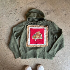 Könnte beinhalten: Ein olivgrüner Kapuzenpullover mit einem quadratischen Aufnäher mit einem Teddybären-Design. Der Aufnäher ist rot mit einem cremefarbenen Rand. Der Pullover ist aus Strickmaterial und hat lange Ärmel.
