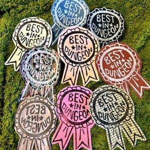 以下が含まれることがあります： 様々な色のリボン型のバッジのコレクションで、円形のデザインの中に「BEST IN DUNGEON」という文字が書かれています。青やピンクなどの色合いのバッジが、緑色の背景に表示されています。