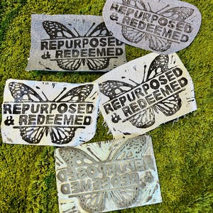 May include: Several fabric patches with a butterfly design and the words "REPURPOSED & REDEEMED" printed in black. The patches are in various shades of gray and white, with a green, textured background.
