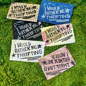 May include: A collection of handmade patches with the text "I WOULD RATHER BE THRIFTING" in various colors and patterns. The patches are on a green, textured surface.