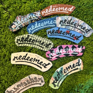 Puede incluir: Un conjunto de parches de tela curvados con la palabra "redeemed" escrita a mano. Los parches presentan diversos patrones, incluyendo diseños florales, a cuadros y geométricos, con una mezcla de colores como azul, rosa y marrón.