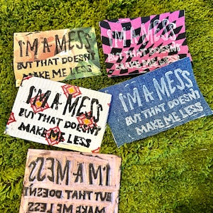 Puede incluir: Cinco impresiones artísticas rectangulares con el texto "I'M A MESS BUT THAT DOESN'T MAKE ME LESS" en varios colores y patrones. Las impresiones están sobre una superficie verde texturizada.
