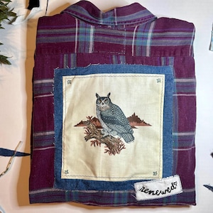 May include: A burgundy plaid shirt, upcycled with a denim-framed patch. The patch displays an owl illustration perched on a branch. The word "renewed" is stitched on a small white label, adding a unique touch to the garment.