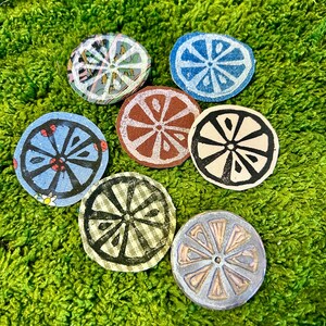 May include: A collection of seven circular coasters, each featuring a unique citrus slice design. The coasters vary in color, including blue, brown, and cream, and are arranged on a bed of green moss. These coasters are ideal for protecting surfaces from drink rings.
