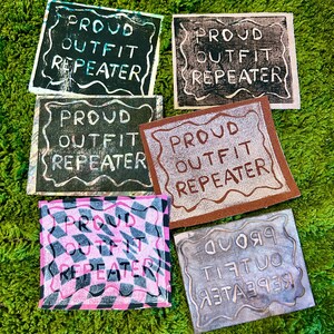 May include: A collection of square art pieces with the text "PROUD OUTFIT REPEATER" in various fonts and colors. The pieces are on a green, textured surface. Some have a black background with white text, while others have pink and black checkered patterns.