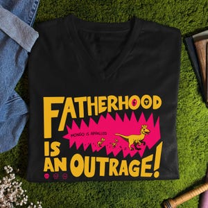 Puede incluir: Camiseta negra con cuello en V con el texto "FATHERHOOD IS AN OUTRAGE!" en amarillo. Un rectángulo rosa dentado contiene un dinosaurio amarillo con una corona y tres dinosaurios más pequeños. También está presente la frase "MONGO IS APPALLED".