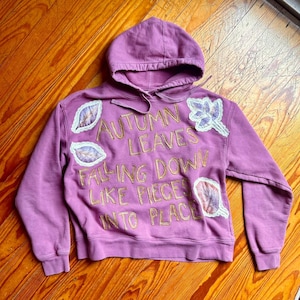 Op de afbeelding: Een mauve hoodie met een trekkoord en lange mouwen. De voorkant heeft gouden geborduurde tekst met de tekst "AUTUMN LEAVES FALLING DOWN LIKE PIECES INTO PLACE", plus bladapplicaties.