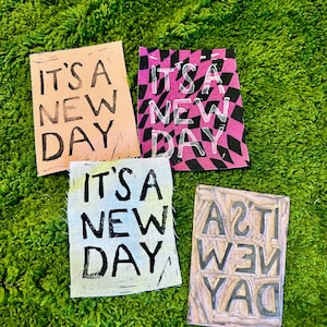 May include: Four rectangular art pieces with the text "IT'S A NEW DAY" in black. The backgrounds are peach, pink checkered, light blue, and a light brown woodblock. The pieces are arranged on a green, textured surface.