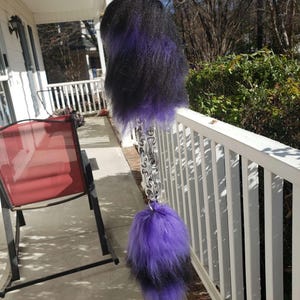 Puede incluir: Un llavero con una cola esponjosa de dos tonos. La parte superior es negra, que pasa a un morado vibrante en la parte inferior. La cola está unida a una cadena plateada y un llavero. El fondo muestra un porche y un entorno al aire libre.