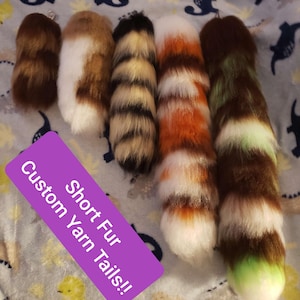 Può includere: Una collezione di sei code corte in finta pelliccia di vari colori, tra cui marrone, bianco, arancione e verde. Le code sono disposte su un tessuto fantasia. Un rettangolo viola con testo bianco recita "Custom Yarn Tails!!" e "Short Fur".