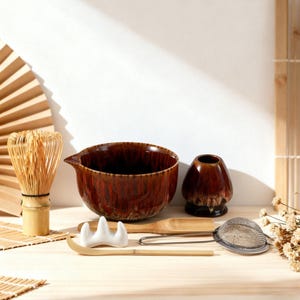 Könnte beinhalten: Ein Matcha-Teeset mit einer braunen Keramikschale, einem Bambusbesen, einer kleinen Keramikvase, einem Bambuslöffel, einem Teesieb und einem Bambuslöffel. Das Set ist auf einer hellen Holzoberfläche mit Bambusmatte und Fächer angeordnet.