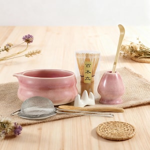 May include: A pink ceramic matcha bowl, a bamboo whisk, a whisk holder, a scoop, a sifter, and a whisk holder. The set includes a bamboo whisk, a scoop, and a small woven coaster. The items are arranged on a wooden surface.