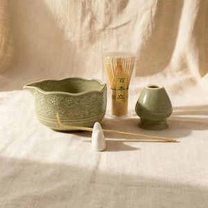 May include: A matcha tea set featuring a green ceramic bowl with floral designs, a bamboo whisk, a whisk holder, and a small ceramic stand. The set is arranged on a neutral-toned fabric.