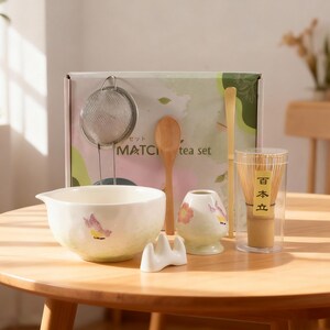Könnte beinhalten: Ein Matcha-Teeset mit einer weißen Keramikschale mit Schmetterlingsmotiven, einem Bambusbesen, einem Holzlöffel, einem Sieb und einem Keramik-Besenhalter. Das Set enthält eine kleine Keramiktasse mit floralen Designs und eine Box mit "MATCH tea set".