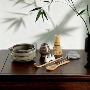 Puede incluir: Un conjunto de herramientas para la ceremonia del té matcha, que incluye un cuenco de cerámica, un batidor de bambú, una cuchara y una cuchara de madera, dispuestos sobre una superficie de madera oscura. El conjunto también incluye un colador de té y un pequeño soporte de cerámica.