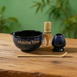 May include: A matcha tea set comprising a navy blue ceramic bowl with "Matcha Time" text, a bamboo whisk, a whisk holder, and a bamboo scoop. The set is displayed on a wooden surface with a green backdrop.