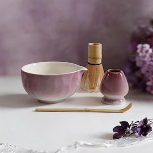 Op de afbeelding: Een keramische matchakom met een mauve en witte gradiëntafwerking, een bamboe garde, een bijpassende mauve keramische gardehouder en een lange, dunne bamboe theeschep. De kom heeft een kleine schenktuit aan één kant en een textuur op het oppervlak.