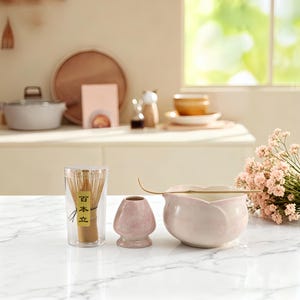 Könnte beinhalten: Ein klarer Behälter mit Bambusbesen und japanischem Text, eine kleine rosa Keramikvase und eine rosa Keramikschale mit einem goldenen Schneebesen. Die Artikel befinden sich auf einer weißen Oberfläche, für eine Teezeremonie.