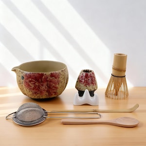 Puede incluir: Un juego de té matcha con un cuenco de cerámica con diseño floral, un batidor de bambú, un soporte para batidor, un colador de acero inoxidable y una cuchara de madera. El juego está dispuesto sobre una superficie de madera clara.