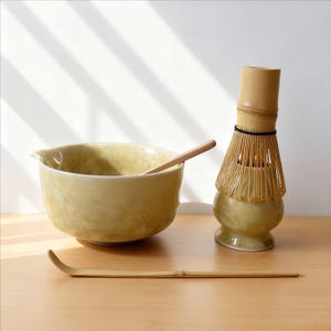 May include: A matcha tea set featuring a pale yellow ceramic bowl with a wooden spoon, a bamboo whisk, and a bamboo scoop. The set is arranged on a light-colored wooden surface, with natural light casting shadows.