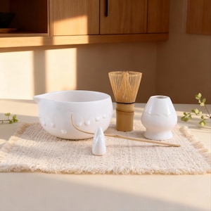 Könnte beinhalten: Ein weißes Matcha-Teeset mit Schale, Bambusbesen, Halter und Löffel. Das Set steht auf einer beigen Webmatte. Im Hintergrund sind Holzschränke und natürliches Licht zu sehen.