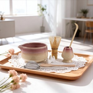 Op de afbeelding: Een matcha theeset op een houten dienblad. De set bevat een keramische kom met een roze en beige gradiënt, een bamboe klopper, een klopperhouder, een lepel, een zeef en een lepel. De lade staat op een wit kanten kleedje.