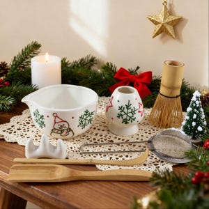 Könnte beinhalten: Ein weihnachtliches Matcha-Teeset mit einer mit Schneemann verzierten Schale, einem Schneebesen und einem Sieb. Das Set enthält einen Bambuslöffel und -halter, einen Keramik-Besenhalter und einen kleinen Weihnachtsbaum. Eine Kerze und ein Sternornament sind ebenfalls vorhanden.