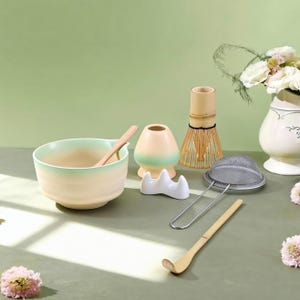 Puede incluir: Un juego de té matcha con un cuenco crema y verde menta, un batidor de bambú, una cuchara y un colador de acero inoxidable. El juego incluye un soporte para batidor de cerámica y un pequeño jarrón con flores blancas.