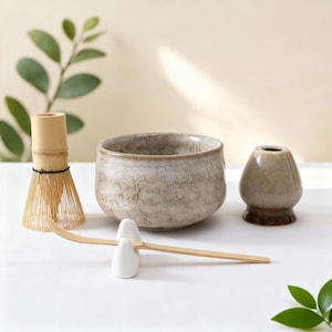 May include: A matcha tea set featuring a ceramic bowl, a bamboo whisk, a whisk holder, and a tea scoop. The bowl has a speckled, off-white glaze. The whisk is light brown, and the whisk holder is white. The set is arranged on a white surface.