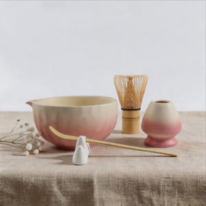 Könnte beinhalten: Ein Matcha-Teeset bestehend aus einer Keramikschale mit rosa-cremefarbenem Farbverlauf, einem Bambusbesen, einem Bambuslöffel auf einem weißen Keramikhalter und einem rosa-cremefarbenen Besenhalter, ergänzt durch einen Zweig weißer Schleierkrautblüten.
