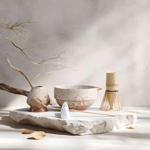 May include: A matcha tea set featuring a ceramic bowl, a bamboo whisk, a small ceramic vase, and a white ceramic spoon. The set is displayed on a white stone surface with a neutral background. Dried branches and leaves add a natural touch.