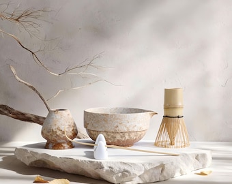 May include: A matcha tea set featuring a ceramic bowl, a bamboo whisk, a small ceramic vase, and a white ceramic spoon. The set is displayed on a white stone surface with a neutral background. Dried branches and leaves add a natural touch.
