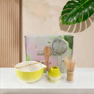 Könnte beinhalten: Ein Matcha-Teeset mit einer gelben Keramikschale, einem Bambusbesen, einem Holzlöffel und einem Edelstahl-Sieb. Das Set enthält einen transparenten Glasbecher mit einem Bambusbesen und einen gelben Keramik-Besenhalter. Auf der Box steht der Text "MAT" und "A tea set".