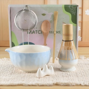 Könnte beinhalten: Ein Matcha-Teeset mit einer hellblauen Keramikschale, einem Bambusbesen, einem Edelstahl-Sieb, einem Holzlöffel und einem Bambuslöffel. Das Set enthält einen weißen Keramik-Besenhalter. Auf der Box steht "Matcha tea set".