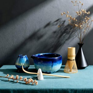 Könnte beinhalten: Ein Matcha-Teeset mit einer blau-weißen Keramikschale, einem Bambusbesen, einem kleinen blauen Keramikbehälter, einem weißen Keramik-Quirlhalter und einer schwarzen Vase. Das Set steht auf einer türkisfarbenen Oberfläche.