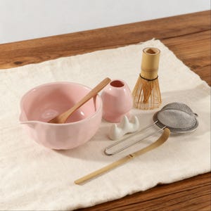 Könnte beinhalten: Ein Set für die Matcha-Teezeremonie, bestehend aus einer rosa Schale, einem Holzlöffel, einem Bambusbesen und einem Teesieb. Die Artikel sind auf einem weißen Tuch angeordnet, ideal für die Teezubereitung.
