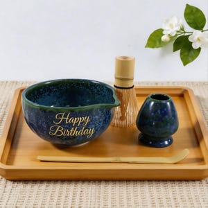 Könnte beinhalten: Ein Matcha-Teeset auf einem Holztablett. Das Set enthält eine blau-grün gesprenkelte Keramikschale mit "Happy Birthday" in goldener Schrift, einen Bambusbesen, einen kleinen Keramikhalter und einen Bambuslöffel.