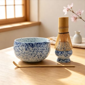 Könnte beinhalten: Eine Matcha-Schale aus Keramik mit blau-weißem Sprenkelmuster, ein Bambusbesen auf einem passenden gesprenkelten Keramikständer und ein langer, dünner, gebogener Bambus-Teelöffel. Das Set zeichnet sich durch ein einheitliches blaues Spritzdesign aus.