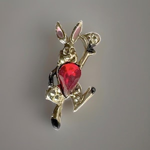 Op de afbeelding: Een goudkleurige metalen broche in de vorm van een rennend konijn. Het konijn is voorzien van een grote, gefacetteerde, peervormige rode kristal in het midden van het lichaam en een kleiner, helder, rond kristalaccent aan de onderzijde.
