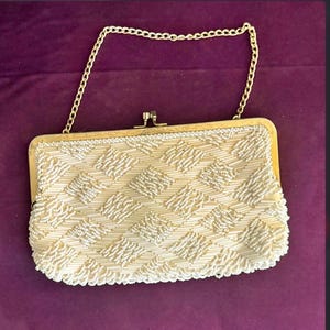 Puede incluir: Bolso de mano con cuentas de color crema con marco y cadena dorados. El bolso rectangular presenta un patrón geométrico de cuentas blancas. El cierre es dorado. Un bolso de noche de estilo vintage.
