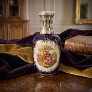 May include: A cobalt blue ceramic fragrance lamp with a metallic silver-toned vented cap. The front features an oval white medallion with a painted scene of two figures in a landscape, surrounded by ornate gold-toned filigree detailing.