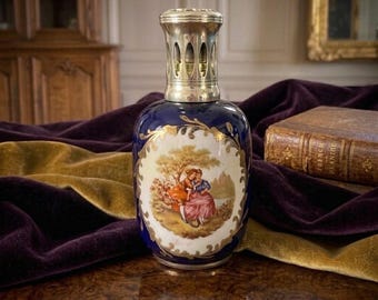 May include: A cobalt blue ceramic fragrance lamp with a metallic silver-toned vented cap. The front features an oval white medallion with a painted scene of two figures in a landscape, surrounded by ornate gold-toned filigree detailing.