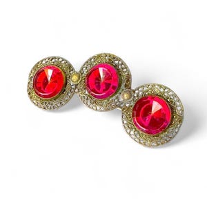 Puede incluir: Broche antiguo con tres grandes gemas rojas redondas y facetadas engastadas en un trabajo de metal calado. El metal tiene un tono dorado y plateado, con pequeños acentos amarillos entre las gemas. El broche se muestra sobre un fondo blanco.