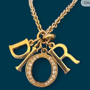 May include: Gold-toned necklace featuring the letters "Dior". The "O" is a circle encrusted with small, clear crystals. The chain and pendant have a shiny finish, set against a dark teal background.