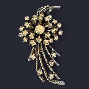Puede incluir: Broche vintage plateado con diseño floral. El broche presenta un gran pedrería transparente central rodeado de pedrería transparente más pequeña. Hebras plateadas retorcidas caen en cascada desde el centro floral, agregando un elemento dinámico.
